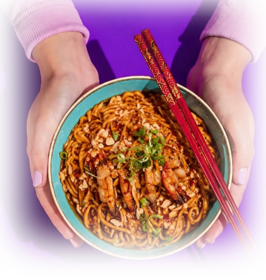 Hands holding a bowl of chili peanut noodles with prawns and red chopsticks, seen from above.